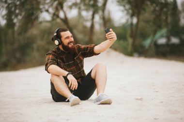 Sakallı, cep telefonu yla mutlu gülümseyen, sahilde işeyleyen ve tatilde selfie fotoğrafı çeken sakallı adam 