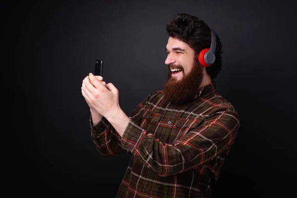 英俊的胡子的家伙戴着耳机 并使用手机在黑暗的背景 — 图库照片