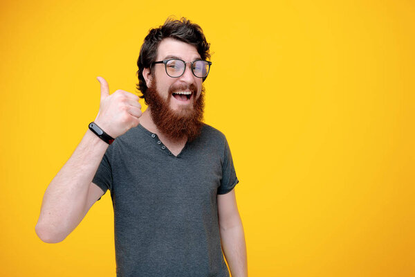 Photo of cheerful bearded guy in glasses, is showing thumb up  over yellow background 
