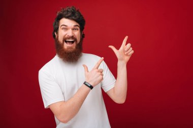 Kırmızı arka plan üzerinden kameraya bakarken fotokopi alanını gösteren sakallı heyecanlı adam 