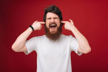Closeup portre genç, kızgın, mutsuz, kırmızı arka plan üzerinde izole kulaklarını kapsayan stresli adam. Olumsuz duygular, yüz ifadeleri. 