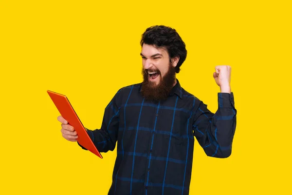 留着胡须的帅哥在黄墙上拿着一块平板电脑 尖叫着 用上升的手臂庆祝 — 图库照片