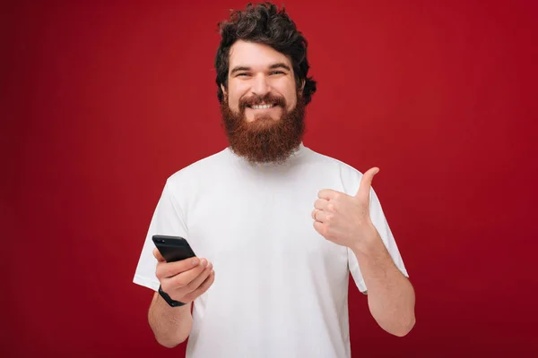 Joven Barbudo Hombre Sobre Pared Roja Sosteniendo Teléfono Inteligente Feliz — Foto de Stock