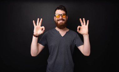 Koyu gri arka plan üzerinde dişli gülümseme ile ok işareti yapıyor güneş gözlüğü takan sakallı bir adamın fotoğrafı 