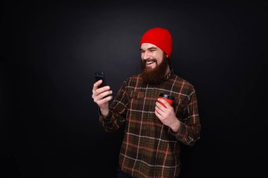 Karanlık duvar üzerinde duran kahve ile fincan tutarken cep telefonu kullanarak sakallı hipster Fotoğrafı