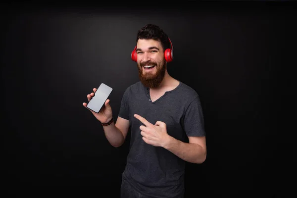 一个留着胡子的男子用耳机指着手机的照片 在黑暗的背景上看着相机 — 图库照片