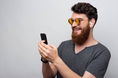 Neşeli sakallı hipster adam akıllı telefon bakıyor ve airpods müzik dinliyor