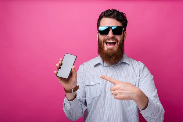 Foto de hombre hipster barbudo alegre en casual apuntando a la pantalla del teléfono inteligente de pie sobre el fondo rosa — Foto de Stock