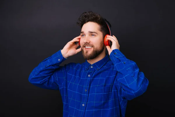 Joven barbudo mirando a un lado escuchando la música a través de un auricular rojo sobre fondo negro . — Foto de Stock