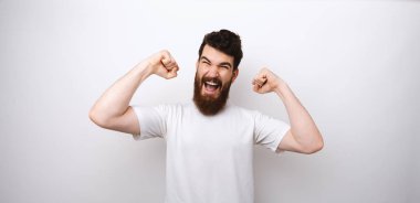 Genç ve çekici sakallı bir adam beyaz arka planda kolları ile kazanan bir jest yapıyor..