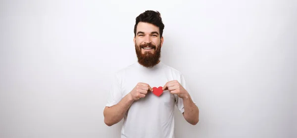 Bärtiger Mann mit einem kleinen roten Papierherz auf weißem T-Shirt an weißer Wand, der in die Kamera lächelt. — Stockfoto