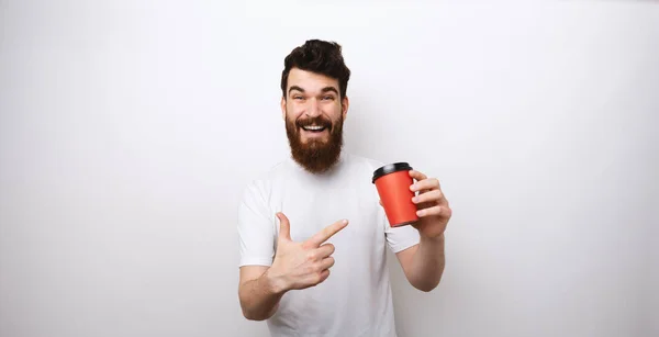 Homme barbu tenant une tasse de café ou de thé en papier rouge et le pointant du doigt sur fond blanc en t-shirt blanc . — Photo