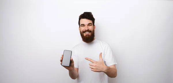 Ung skäggig man som visar och pekar på sin telefon på vit bakgrund. — Stockfoto