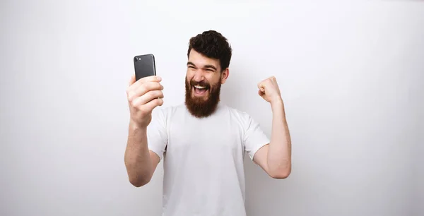 Homem barbudo fazendo um gesto vencedor e olhando para seu telefone no fundo branco . — Fotografia de Stock