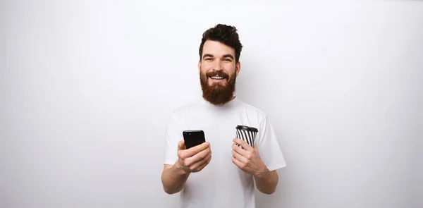 Молодой бородатый мужчина в белой рубашке на белом фоне держит свой телефон и чашку кофе или чая забрать или пойти . — стоковое фото