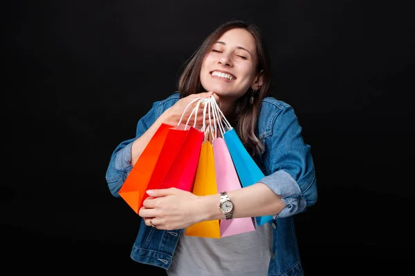 Ung kvinna omfamna sina shoppingväskor och njuta av dem med slutna ögon på svart bakgrund. — Stockfoto