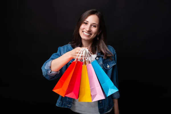 Όμορφη νεαρή γυναίκα χαμογελά στην κάμερα που δείχνει πολύχρωμες σακούλες για ψώνια σε μαύρο φόντο. — Φωτογραφία Αρχείου