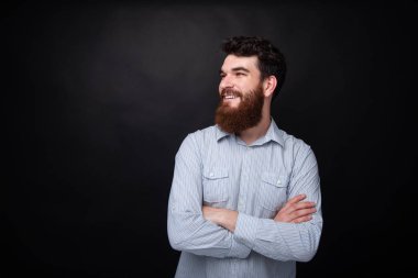 Koyu gri arka plan üzerinden uzağa bakan, çapraz elleri ile sakallı adam portresi 