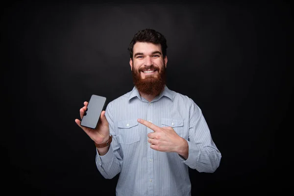 Foto de homem barbudo alegre apontando para spartphone enquanto olha e sorrindo para a câmera — Fotografia de Stock