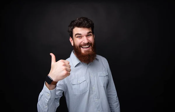 留着胡须的男子在孤立的黑暗背景上竖起大拇指，带着牙齿的微笑的肖像 — 图库照片