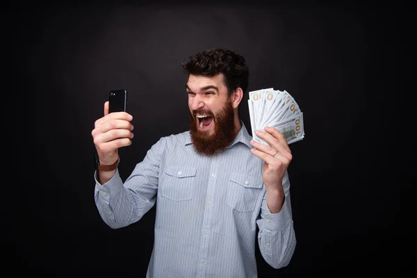 Handsome excited bearded guy, screaming and celebrating his won, while holding a mobile phone and a lot of money — Stock Photo, Image