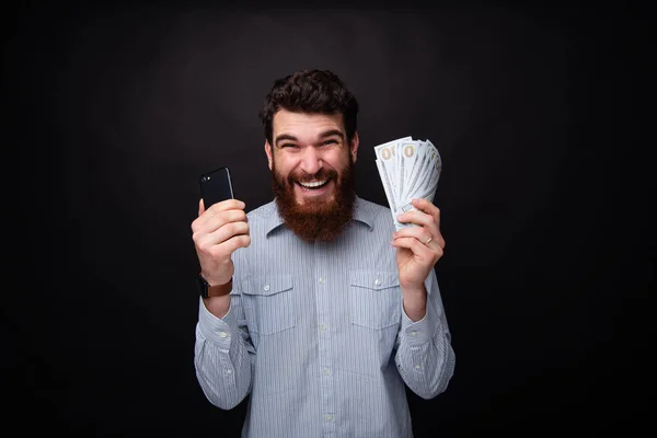 兴奋的胡子的家伙的照片，拿着智能手机和很多钱，庆祝他赢得了德拉克孤立的背景 — 图库照片