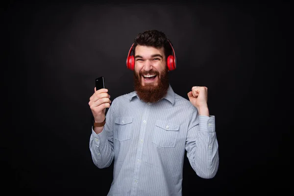 Foto de un joven barbudo alegre, celebrando con los brazos levantados, mientras usa un teléfono móvil y auriculares —  Fotos de Stock