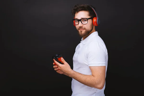 Foto de un joven guapo, mirando a la cámara, sosteniendo una taza de café, usando teléfonos móviles y auriculares — Foto de Stock