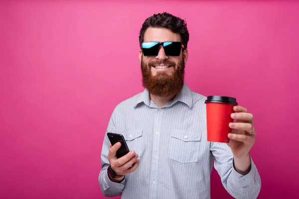 Jovem barbudo alegre segurando um copo take away e seu telefone no fundo rosa . — Fotografia de Stock