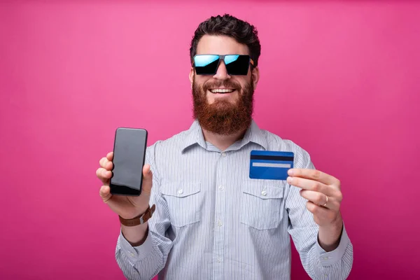 Hombre barbudo con gafas de sol está mostrando a la cámara su teléfono con pantalla en blanco y tarjeta de crédito sobre fondo rosa . — Foto de Stock