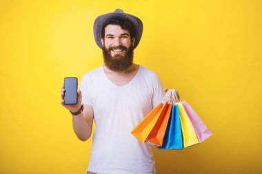 Mutlu genç sakallı adam telefon ekranı sarı arka plan üzerinde bazı alışveriş çantaları tutuyor gösterir.