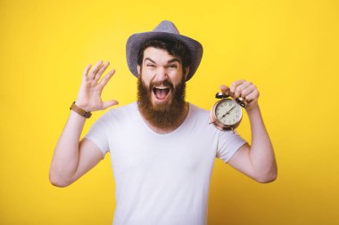 excited young bearded man wearing a summer hat is screaming, shouting or shocked about the time. He is holding an alarm clock. clipart