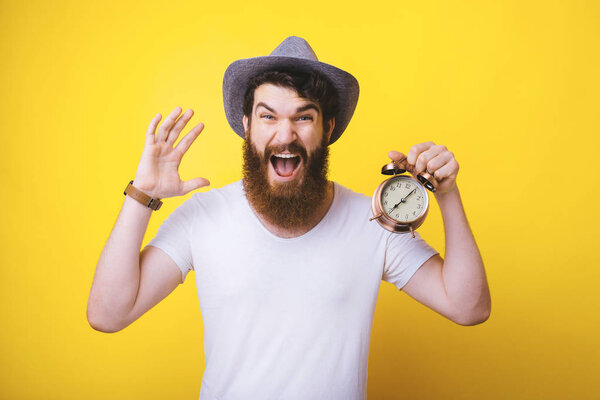 excited young bearded man wearing a summer hat is screaming, shouting or shocked about the time. He is holding an alarm clock.