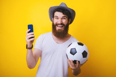 Heyecanlı bir adam elinde bir futbol topu ve akıllı telefonu tutuyor. Futbol maçı izlemek, online mobil akış. Spor bahisleri.