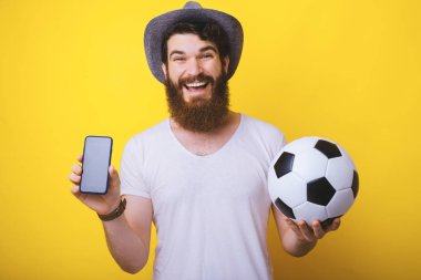 Heyecanlı sakallı adam bir futbol topu ve akıllı telefon tutan kameraya bakıyor. 