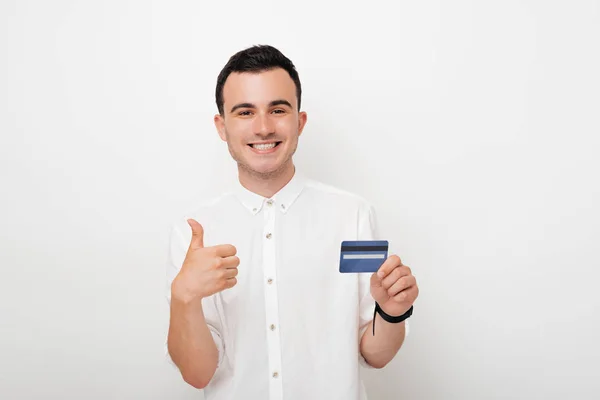 Joven alegre mostrando gesto de pulgar hacia arriba y con otra mano sosteniendo una tarjeta de crédito sobre fondo blanco —  Fotos de Stock