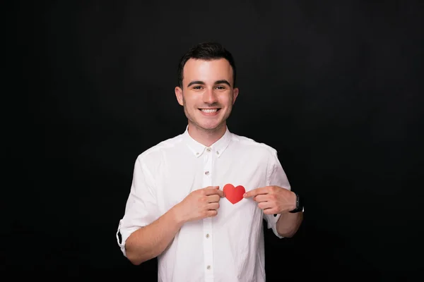 Joven alegre sosteniendo con sus manos un corazón de papel rojo sobre fondo negro —  Fotos de Stock