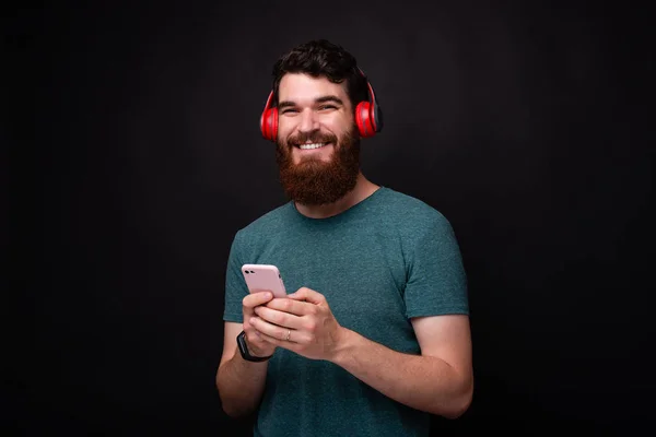 Foto de hombre con barba mirando a la cámara y sonriendo, hombre sosteniendo el teléfono inteligente y usando auriculares rojos — Foto de Stock