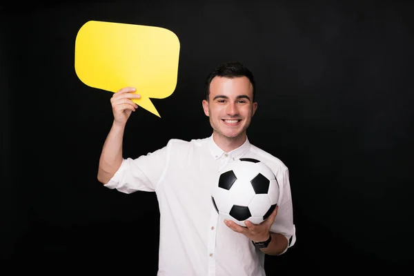 Jovem alegre no fundo preto segurando uma bola de futebol ou futebol e uma bolha de fala amarela. Pensamentos sobre esportes . — Fotografia de Stock