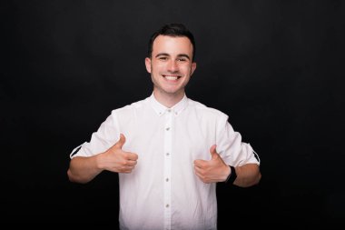 Beyaz gömlekli genç mutlu adam başparmak gösterir ve siyah bir arka plan üzerinde kameraya gülümseyerek.