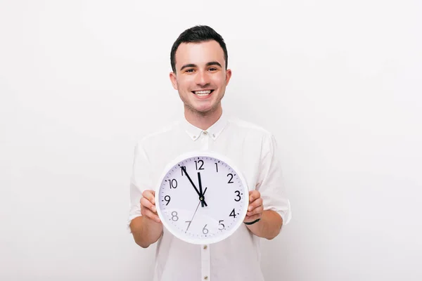 Ein glücklicher junger Mann hält eine weiße Uhr in der Hand und blickt in die Kamera im Studio auf weißem Hintergrund. — Stockfoto