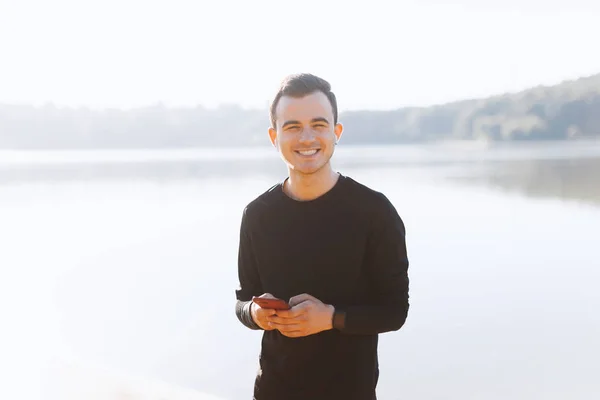 Giovane gioioso sta sorridendo alla fotocamera un tenendo un telefono nel parco . — Foto Stock