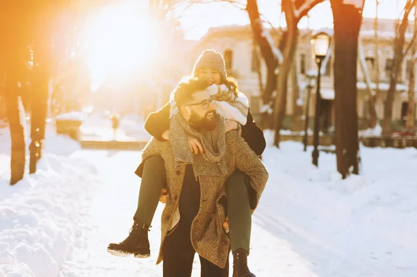 Joven pareja alegre, donde el hombre está sosteniendo a su chica en su espalda, ambiente romántico de invierno . — Foto de Stock
