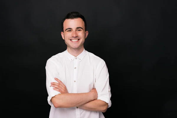 Joven alegre con camisa blanca sonríe a la cámara sobre fondo negro cruzando brazos . —  Fotos de Stock