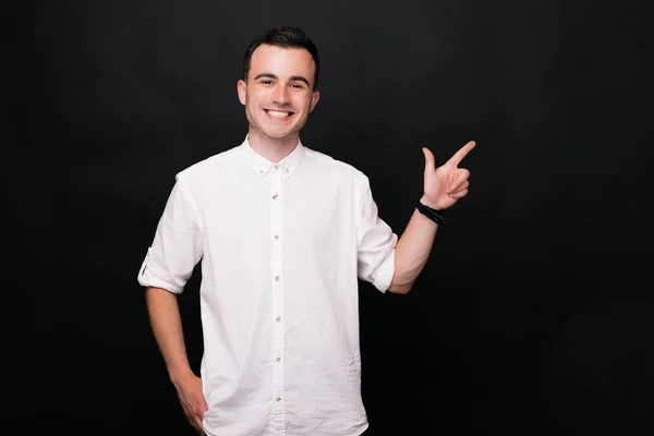 Jovem homem alegre em camisa branca está mostrando com a mão no fundo preto . — Fotografia de Stock