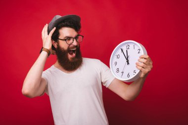 Genç sakallı hipster beyaz bir saat tutan ve şok izlerken, çığlık ve kırmızı arka plan üzerinde vurguladı.