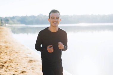 Gün doğumunda sahilde koşan kulaklık takan genç adam kameraya gülümseyerek.