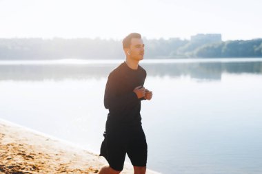 Plajda göl veya deniz kenarında koşan konsantre genç adam güneş ışığında spor kıyafetler giyiyor.