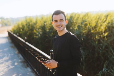 Genç gülümseyen sporcu dışarıda durmuş akıllı telefonuna bir şeyler yazıyor, kenara bakıyor. Sabah temiz hava solunumu.