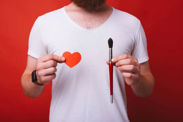 I love painting! Man wearing white shirt is holding a red heart shaped paper and a red painting brush on red background.